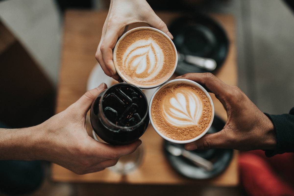 Três pessoas brindando com diferentes bebidas de café, incluindo latte art e café gelado