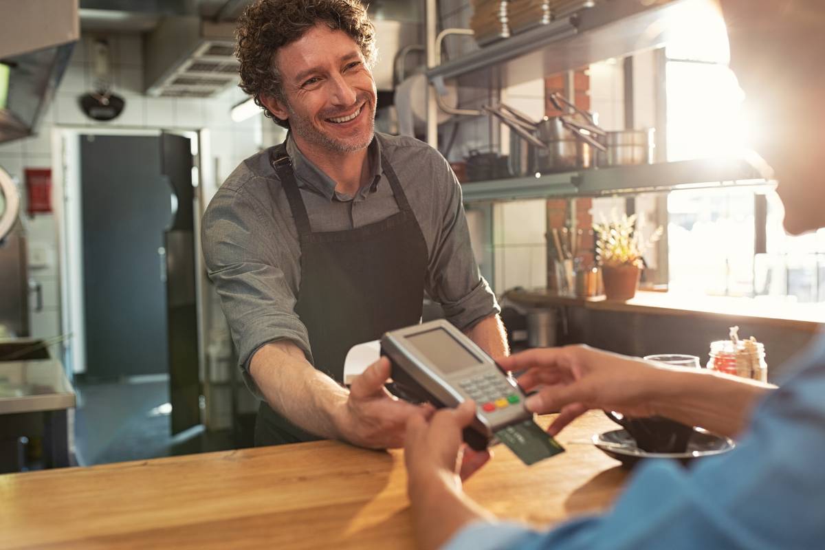 Homem sorridente com avental em um restaurante, segurando uma maquininha de cartão enquanto um cliente faz um pagamento eletrônico. Ambiente iluminado com cozinha ao fundo.