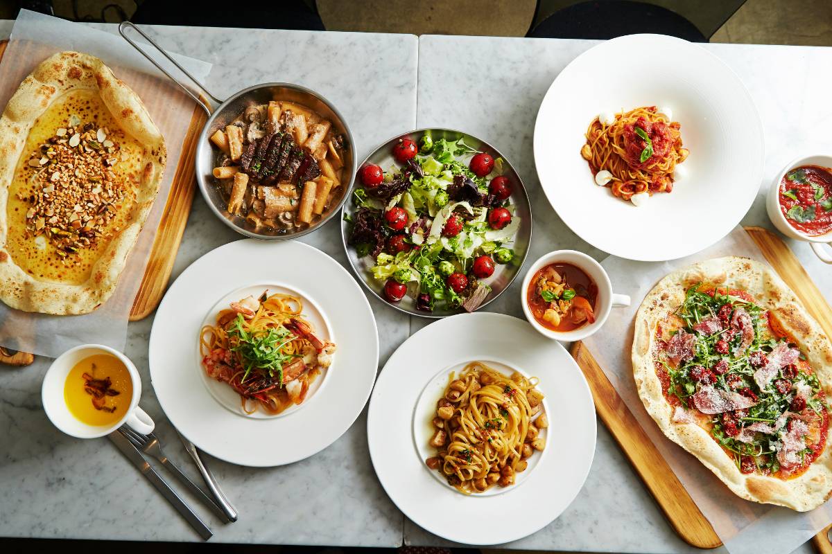 Mesa com diversos pratos italianos, incluindo massas, salada, e pizza, organizados de forma elegante.