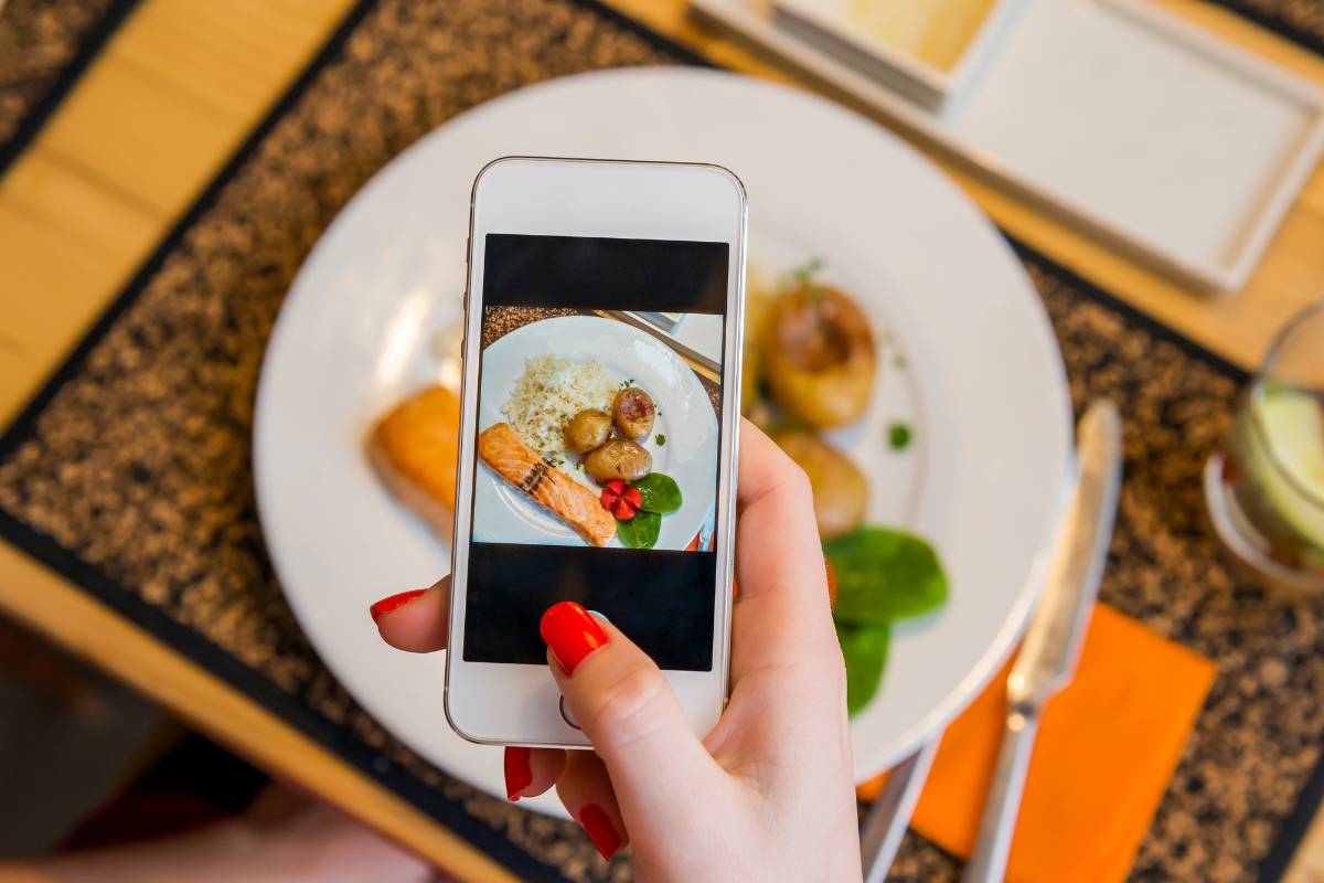 Mulher tirando foto de um prato de salmão grelhado com batatas e arroz usando um smartphone em restaurante.