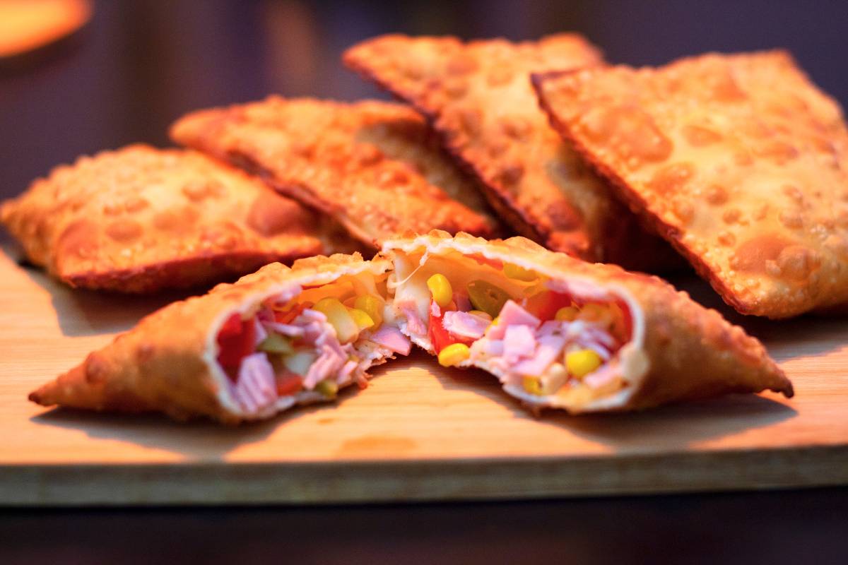 Pastéis crocantes em uma tábua de madeira, com um pastel aberto revelando recheio colorido de presunto, milho, tomate e queijo