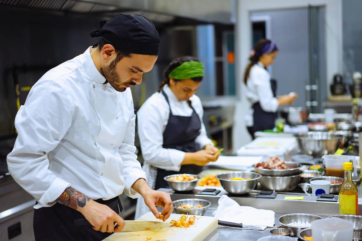 Cozinheiros trabalhando em equipe em uma cozinha profissional, cortando e preparando alimentos com utensílios organizados.