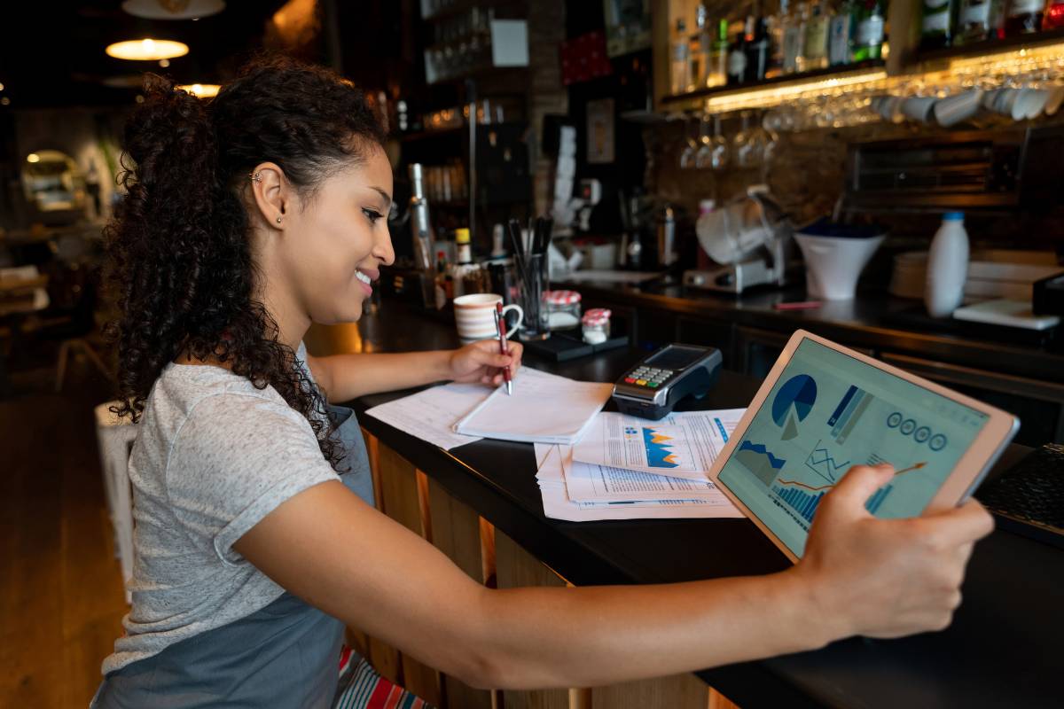 Funcionária de restaurante revisando gráficos financeiros em um tablet e fazendo anotações em um caderno, em um ambiente de bar com diversos utensílios ao fundo.