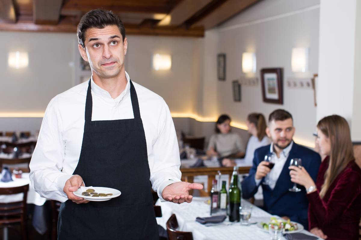 Garçom segurando um prato com algumas moedas, olhando para frente com expressão incerta, enquanto clientes conversam em uma mesa ao fundo em um restaurante.