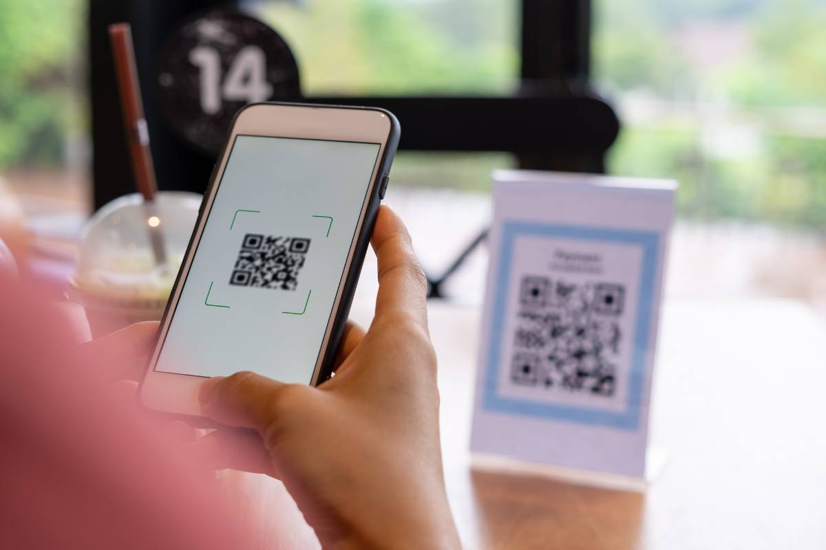 Pessoa usando smartphone para escanear QR Code em um restaurante, com o código exibido em um suporte na mesa.