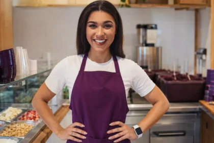 Proprietária sorridente de avental roxo posa em uma loja de açaí com balcão de frutas, granola e outros complementos.