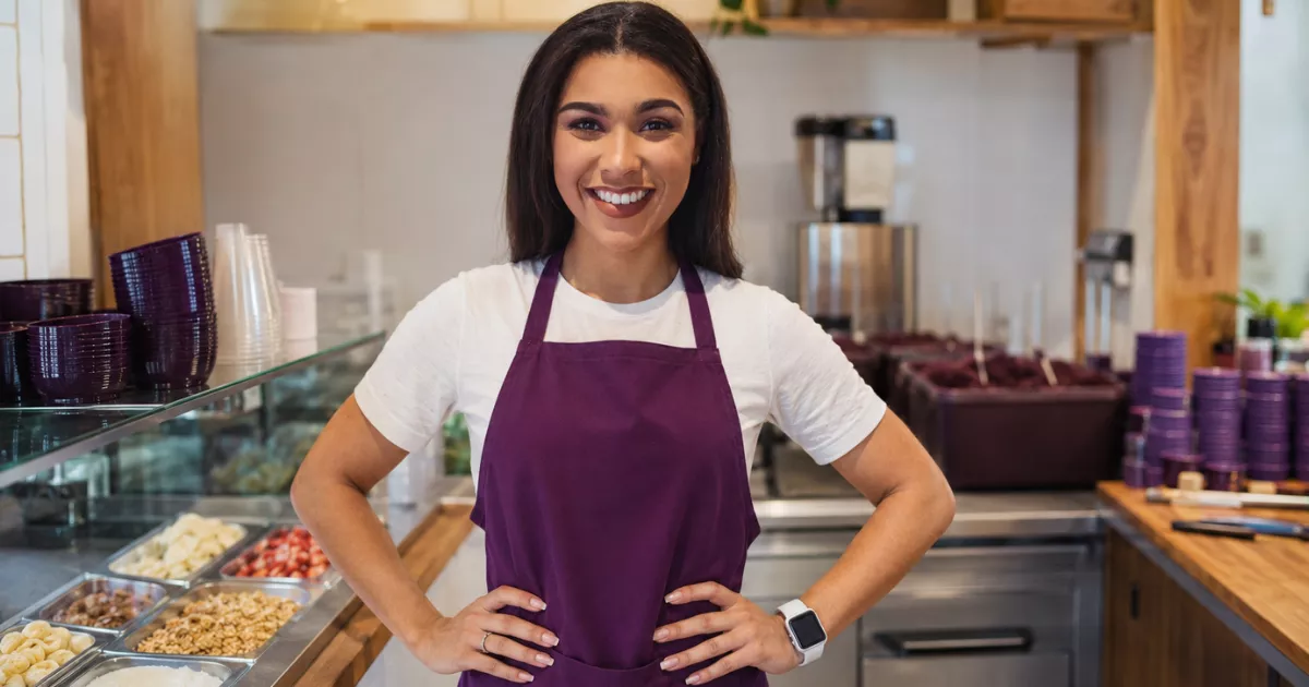 Proprietária sorridente de avental roxo posa em uma loja de açaí com balcão de frutas, granola e outros complementos.