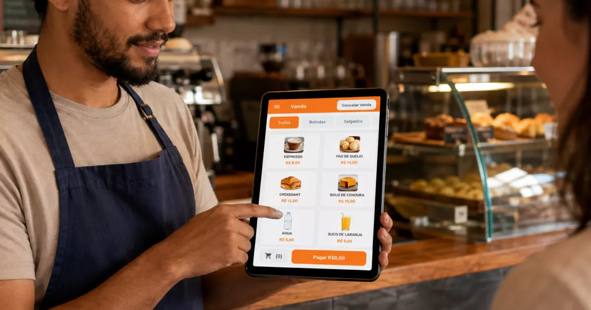 Garçom de cafeteria registra pedido em tablet com tela do ControleNaMão, diante do balcão e da vitrine de salgados.