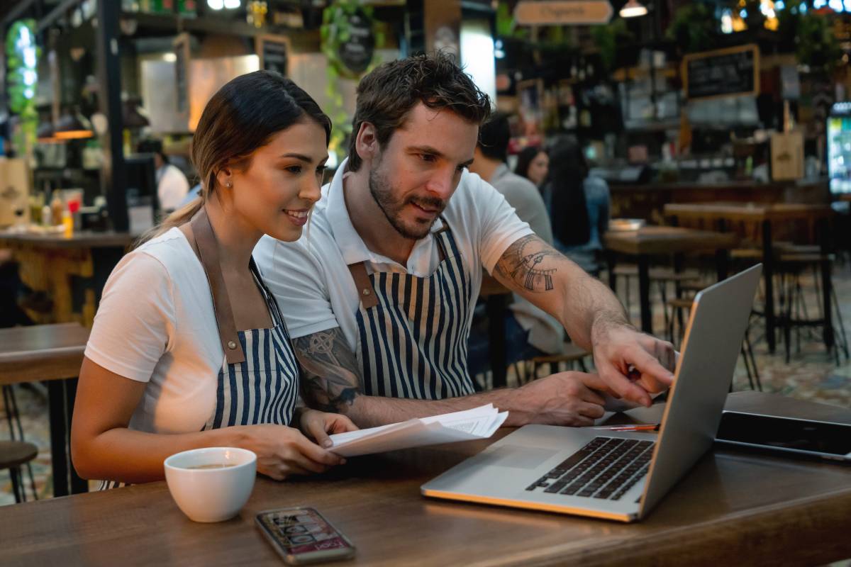 Dois donos de restaurante, vestindo aventais, analisando dados financeiros em um laptop dentro do estabelecimento, demonstrando foco e trabalho colaborativo.