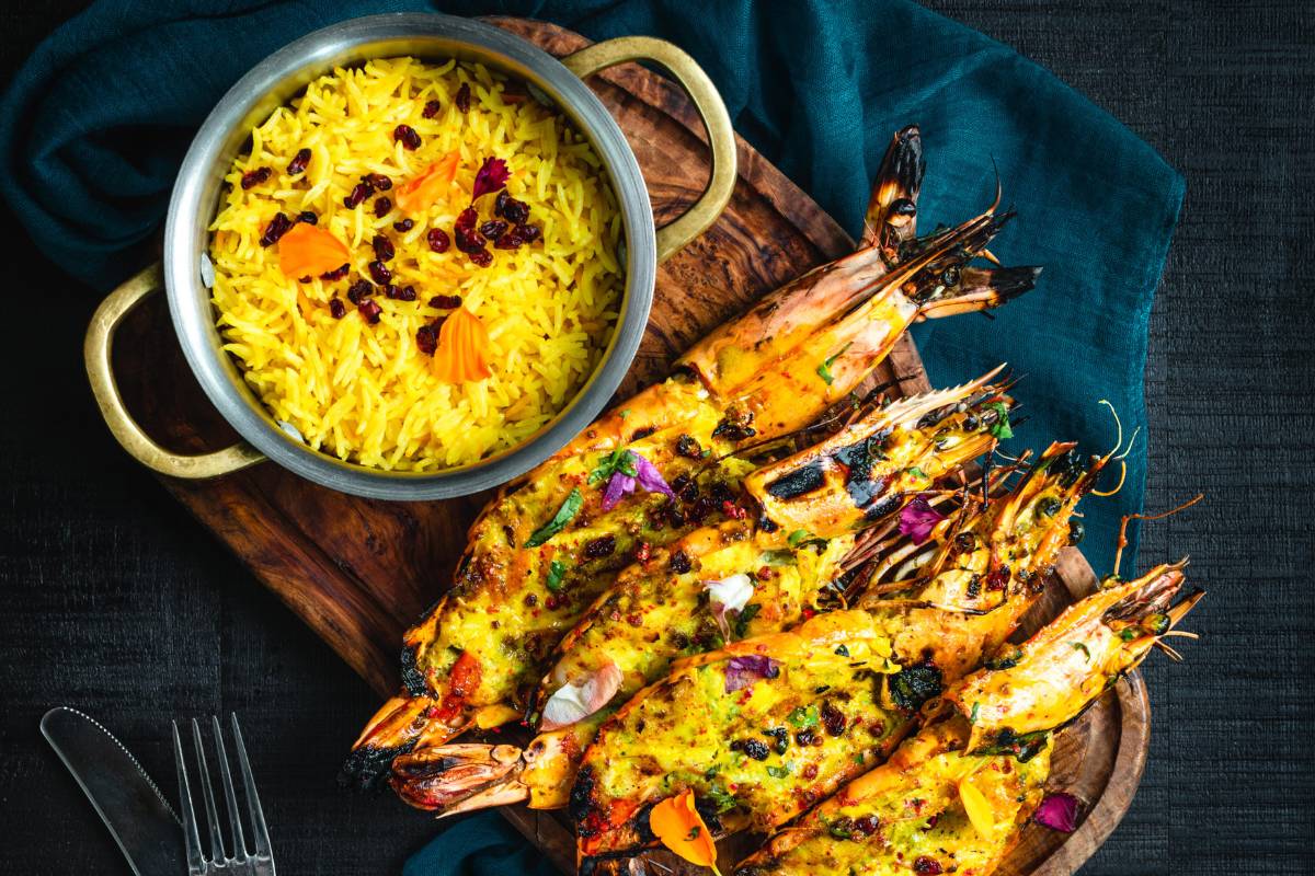 Tigela de arroz amarelo com açafrão e camarões grelhados em uma tábua de madeira, decorada com flores e ervas