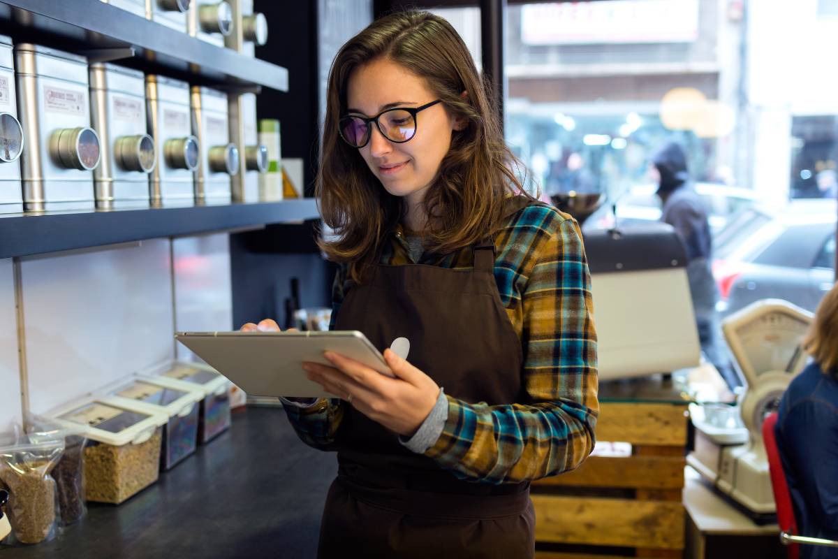 Funcionária de cafeteria analisando dados em um tablet em um ambiente repleto de produtos e insumos organizados.