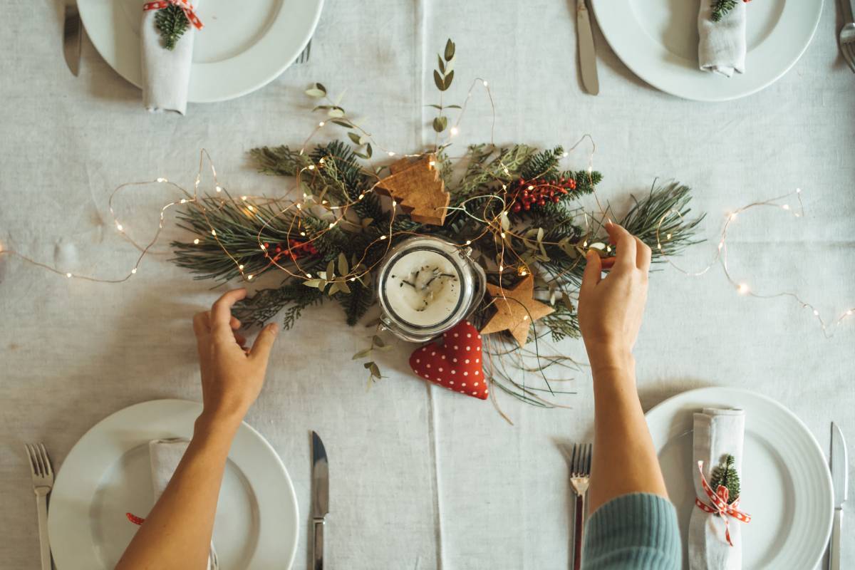 Uma mesa de jantar com decoração natalina, incluindo um centro de mesa com ramos verdes, luzes de LED, uma vela e enfeites festivos. Guardanapos decorados complementam a ambientação aconchegante e festiva.