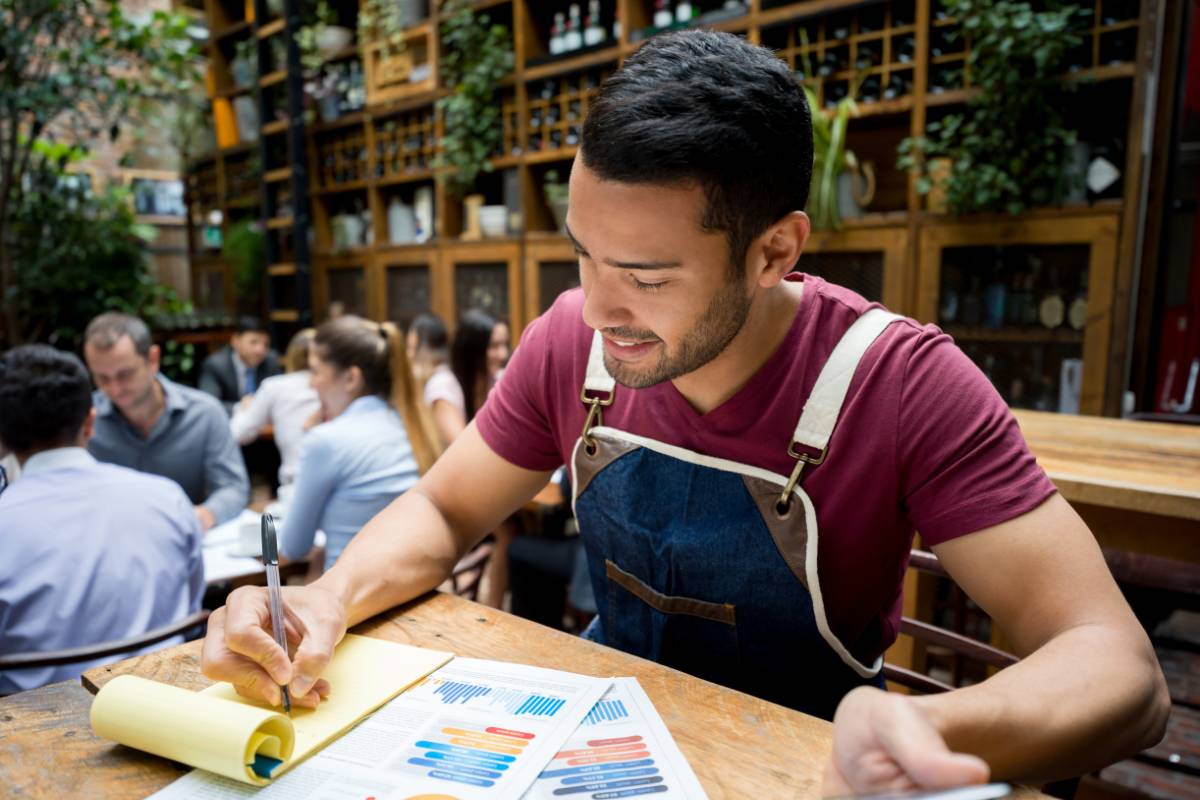 Gerente de restaurante preenchendo documentos em uma mesa ao ar livre, com gráficos e tabelas no papel.