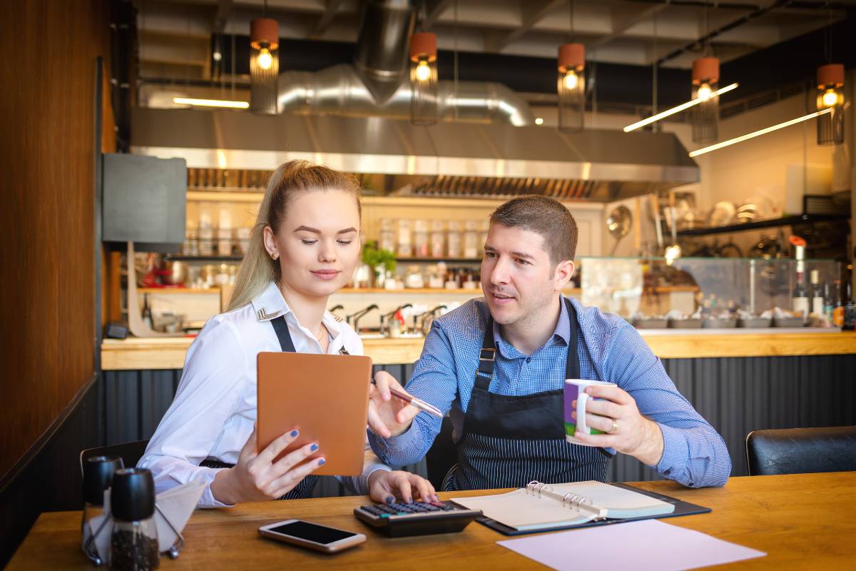 Um homem e uma mulher, também gestores de restaurante, revisando informações em um tablet enquanto discutem estratégias financeiras em um ambiente moderno de restaurante.