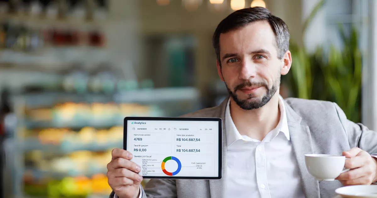 Gestor em cafeteria segura um tablet com relatório de vendas enquanto toma café.