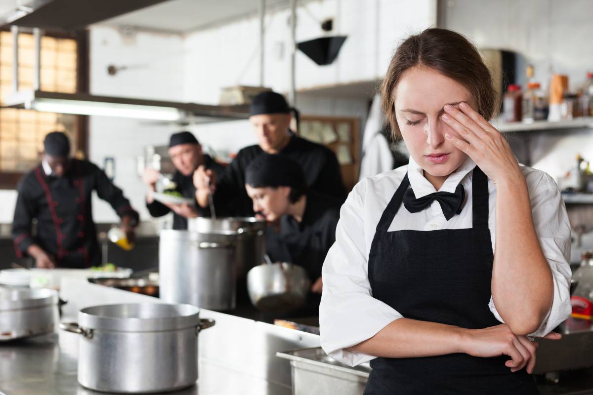 Garçonete com expressão cansada em uma cozinha movimentada, ilustrando a sobrecarga de trabalho e a necessidade de calcular corretamente as horas extras em bares e restaurantes