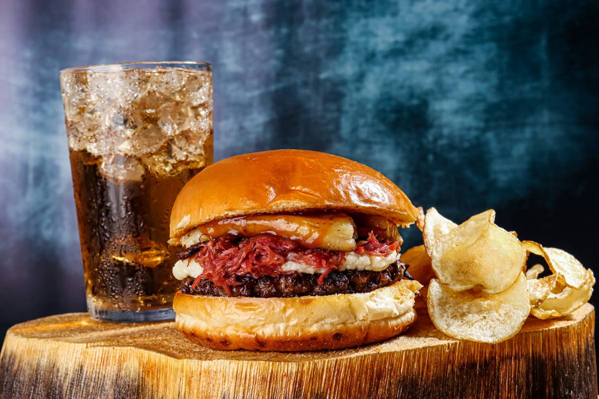 Hambúrguer artesanal com carne desfiada, queijo, e chips ao lado, acompanhado de um copo de refrigerante com gelo.