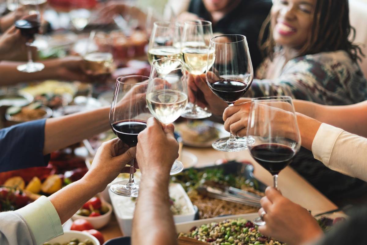 Pessoas brindando com taças de vinho em uma refeição de grupo, com pratos diversos ao fundo.