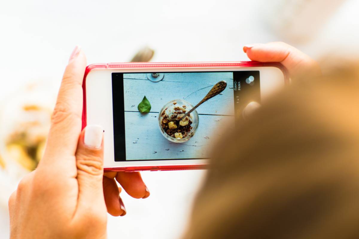 Mão segurando um celular para fotografar uma sobremesa com folhas e colheres decorativas sobre mesa branca.