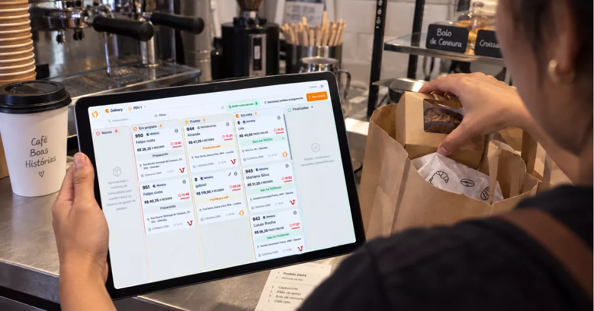 Funcionária de cafeteria confere pedidos na Central de Delivery do ControleNaMão enquanto organiza cafés e doces em embalagens para entrega.