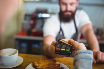 Cliente aproxima cartão de uma maquininha em balcão de cafeteria, enquanto atendente recebe o pagamento durante atendimento em restaurante.