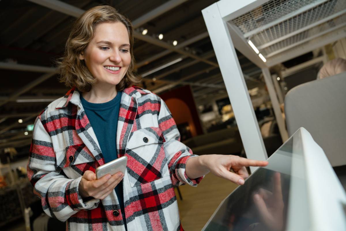 Cliente sorrindo enquanto utiliza totem de autoatendimento - Mulher sorridente usando camisa xadrez interage com um totem de autoatendimento, segurando um celular enquanto faz um pedido ou consulta informações