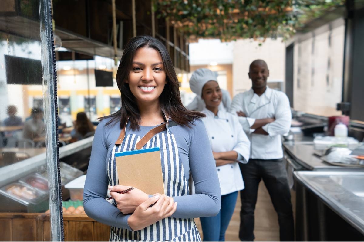 Proprietária de restaurante sorrindo com um bloco de notas nas mãos, acompanhada por sua equipe de cozinha ao fundo.