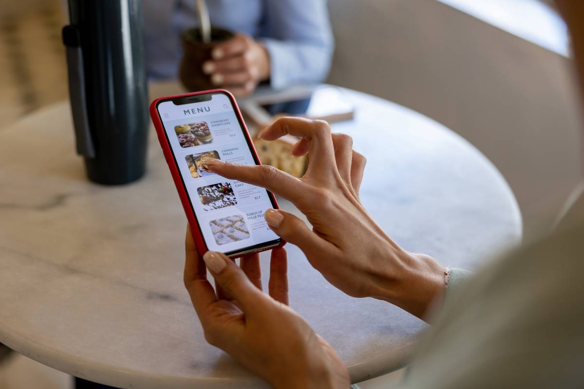 Pessoa utilizando um menu digital em um smartphone em um restaurante, escolhendo itens diretamente pelo aplicativo.