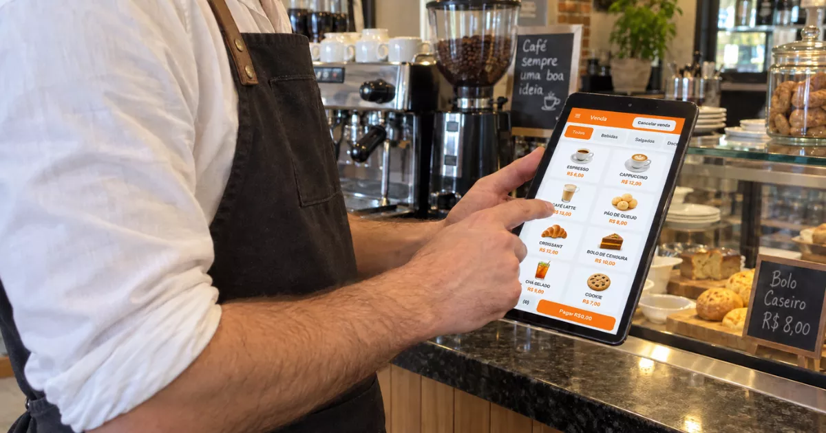 Atendente de cafeteria registra pedido em tablet com o PDV mobile do ControleNaMão diante da máquina de espresso e vitrine de salgados.