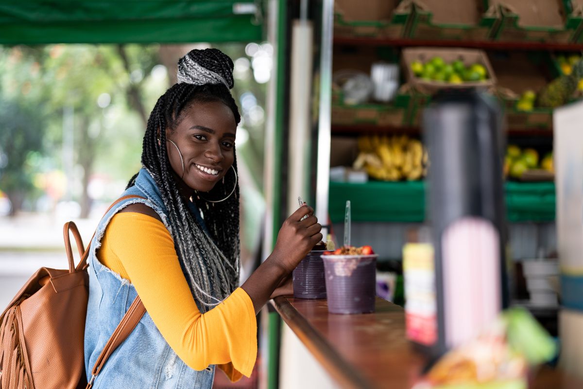 Mulher sorrindo ao lado de um balcão com potes de açaí, em uma barraca com frutas expostas ao fundo.