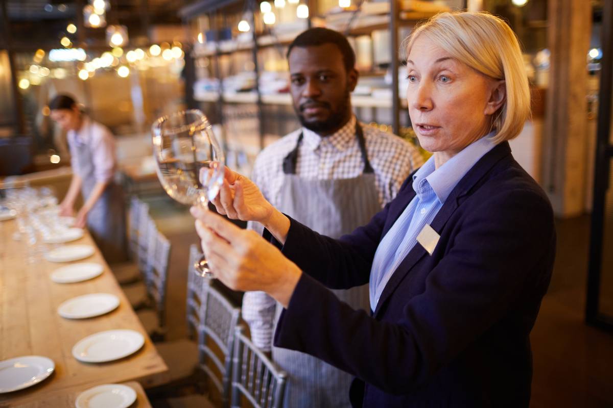 Gerente de restaurante inspecionando uma taça com a equipe ao fundo, durante o processo de preparação do estabelecimento.