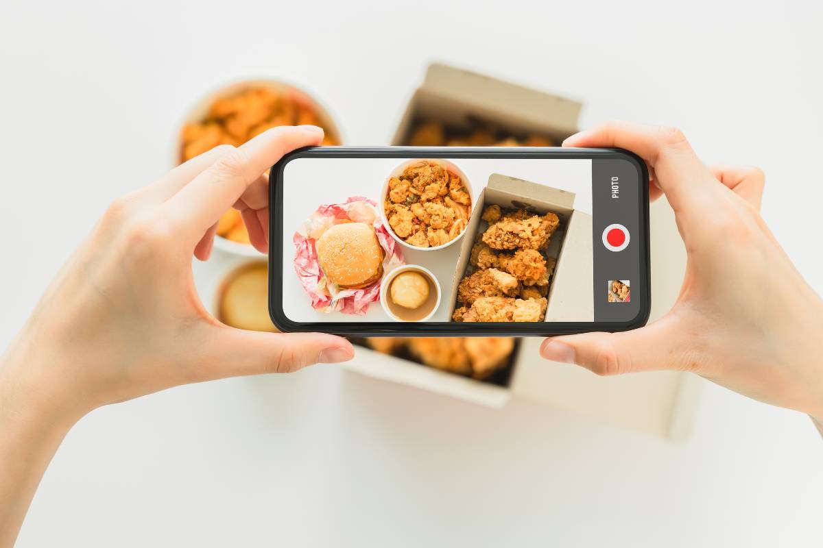 Mãos segurando um smartphone fotografando uma refeição com hambúrguer e frango empanado em embalagens.