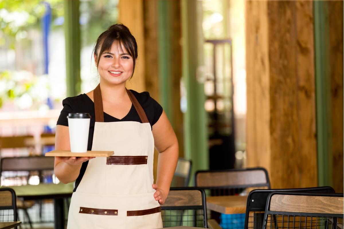 Garçonete de cafeteria sorrindo enquanto segura uma bandeja com uma bebida para servir em um espaço decorado com madeira e plantas.