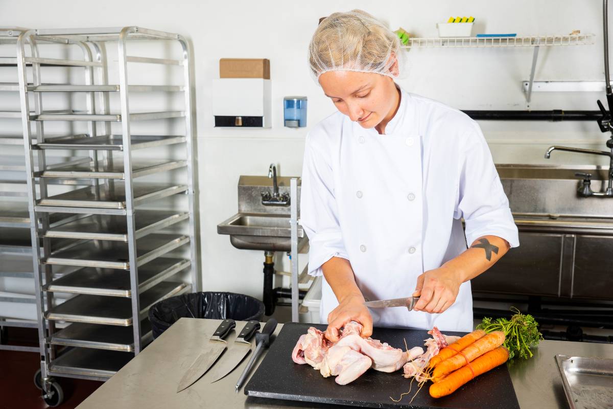 Chef manipulando frango cru em uma cozinha profissional com utensílios e vegetais na bancada.