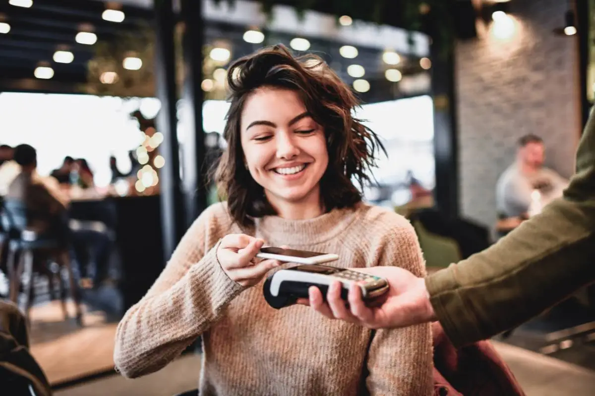 Mulher sorrindo enquanto realiza pagamento por aproximação com smartphone em uma maquininha de cartão em um restaurante.