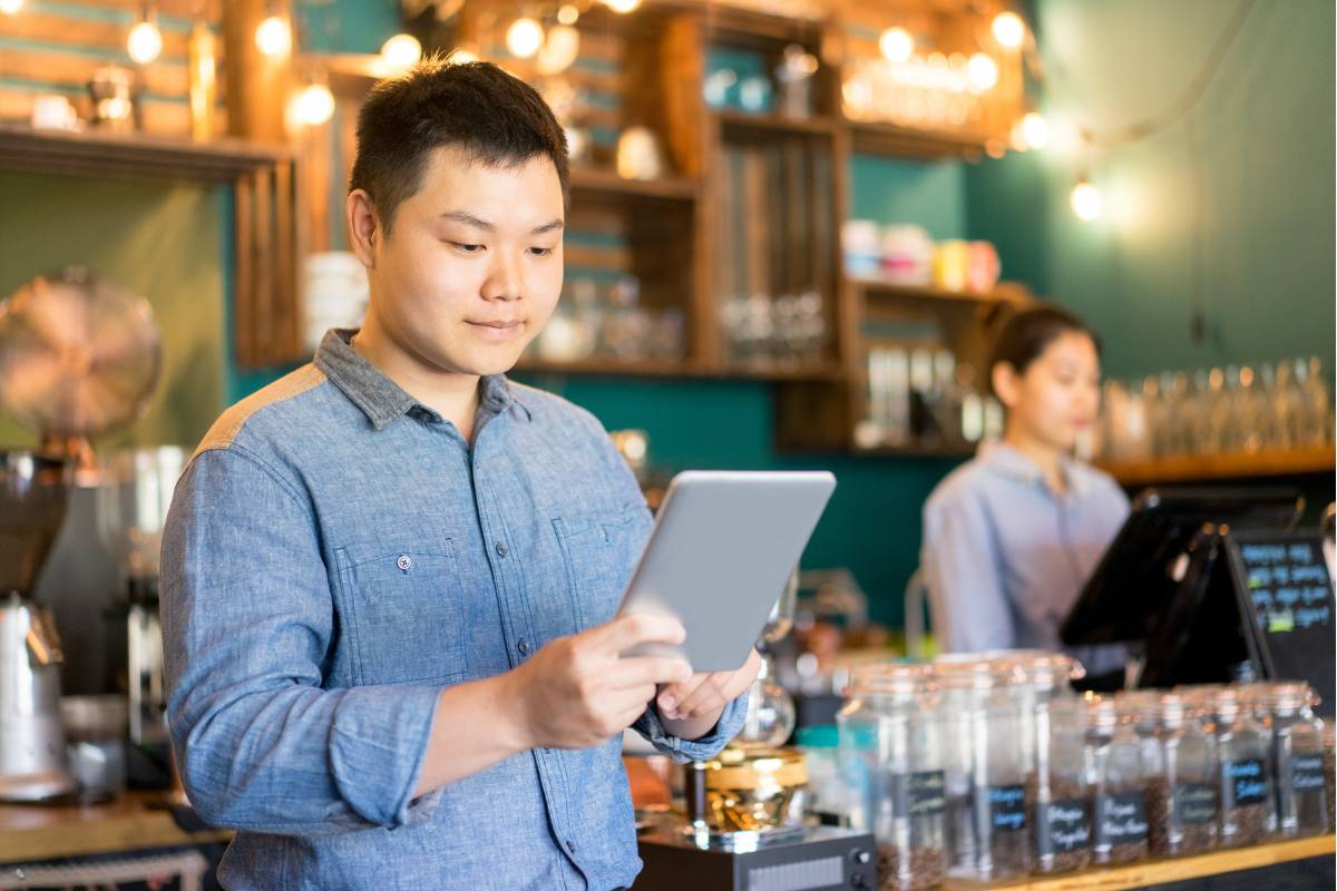 Barista utilizando um tablet para gerenciar pedidos em uma cafeteria moderna, com decoração aconchegante ao fundo.