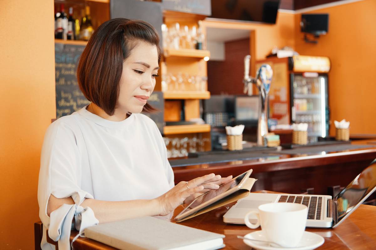 Mulher usando um tablet e laptop para gerenciar um bar, sentada em uma mesa com uma xícara de café ao lado