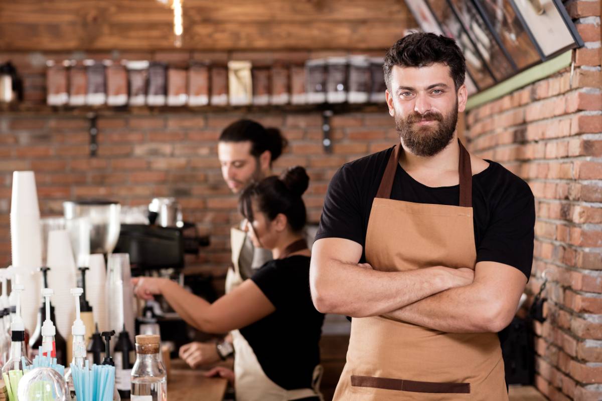 Barista confiante à frente do balcão de uma cafeteria, com equipe preparando bebidas ao fundo.