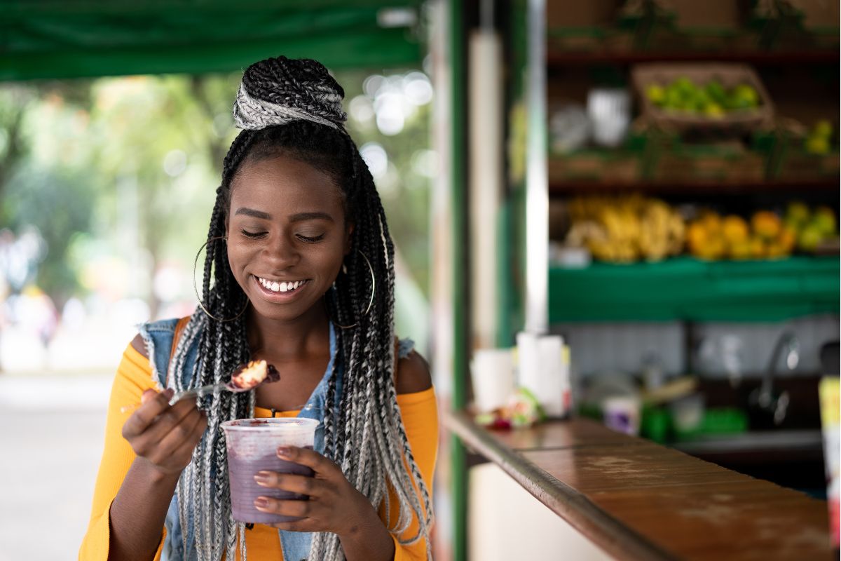 Mulher sorrindo enquanto saboreia um pote de açaí em uma barraca ao ar livre.