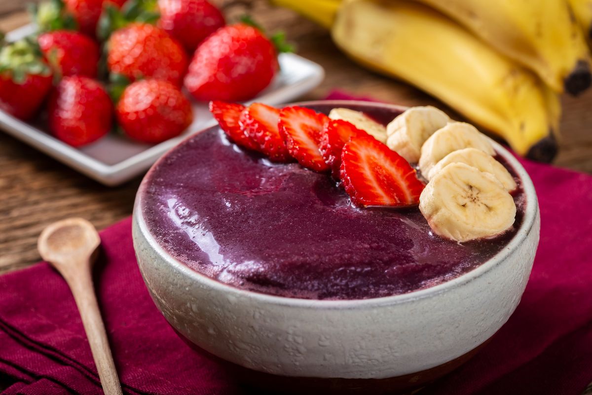 Tigela de açaí com fatias de morango e banana, acompanhada por frutas frescas ao fundo.