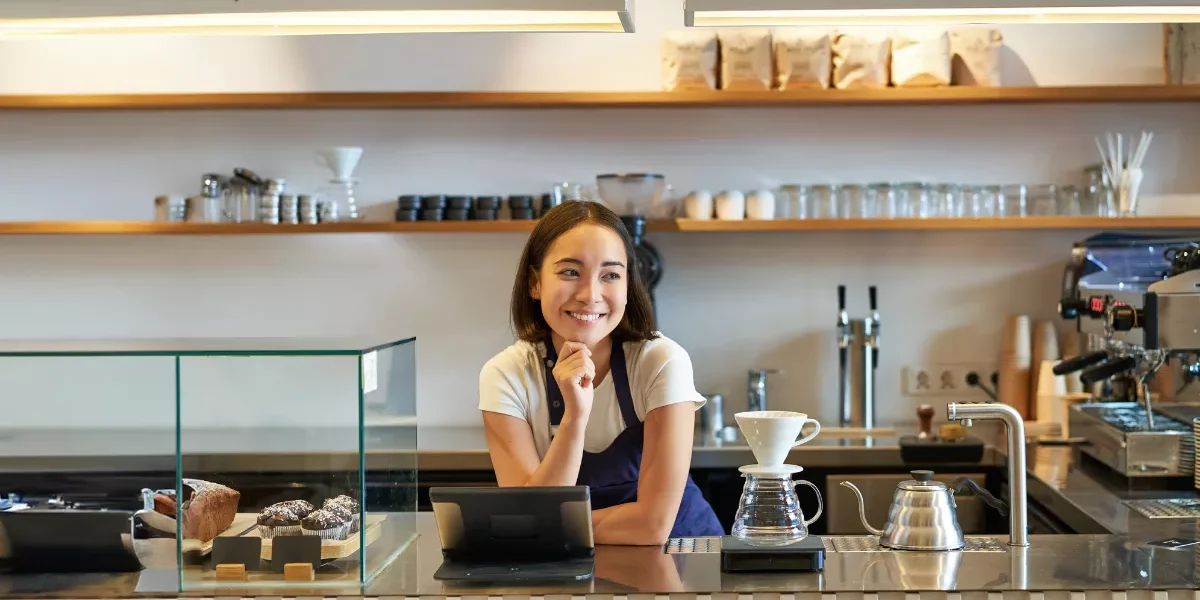Barista sorridente atrás do balcão de cafeteria pequena, com tablet, máquina de espresso, doces expostos e equipamentos para café filtrado.