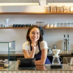 Barista sorridente atrás do balcão de cafeteria pequena, com tablet, máquina de espresso, doces expostos e equipamentos para café filtrado.