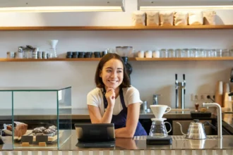 Barista sorridente atrás do balcão de cafeteria pequena, com tablet, máquina de espresso, doces expostos e equipamentos para café filtrado.