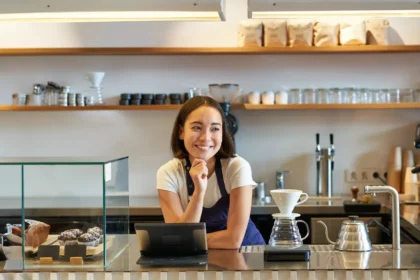 Barista sorridente atrás do balcão de cafeteria pequena, com tablet, máquina de espresso, doces expostos e equipamentos para café filtrado.