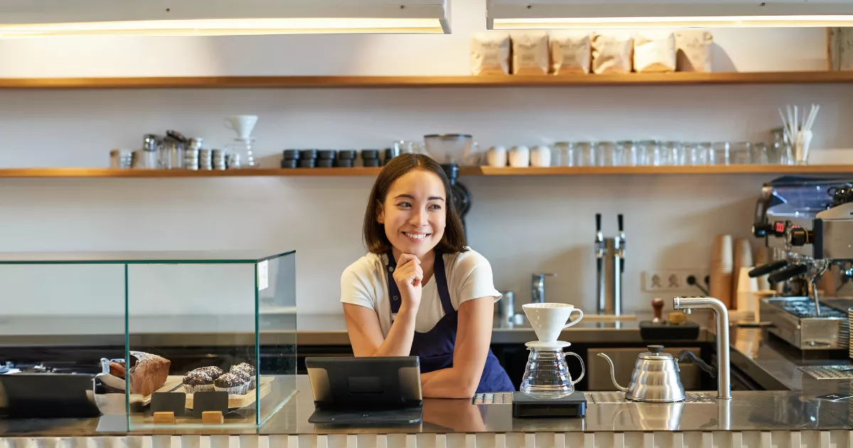 Barista sorridente atrás do balcão de cafeteria pequena, com tablet, máquina de espresso, doces expostos e equipamentos para café filtrado.