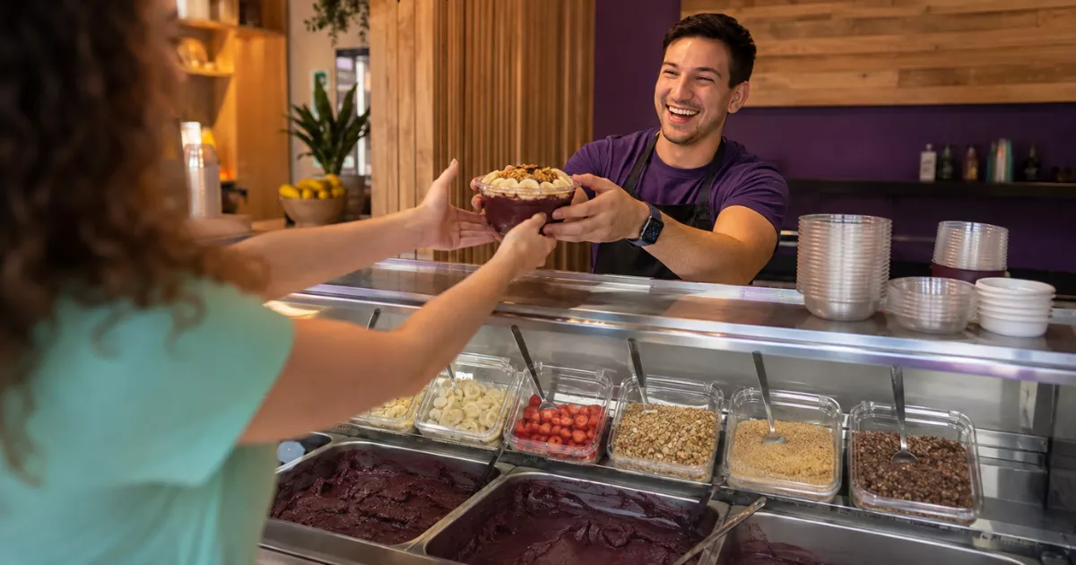 Atendente sorridente entrega uma tigela de açaí com banana e granola para cliente no balcão de uma loja de açaí brasileira.