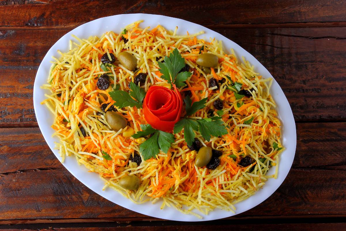 Salpicão decorado com batata palha, passas, azeitonas e uma flor de tomate, servido em uma travessa branca.