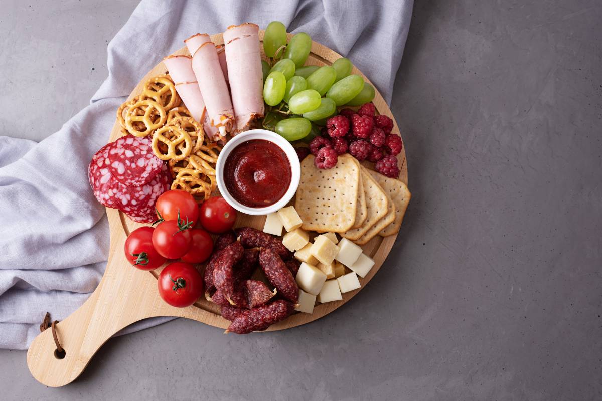 Tábua de frios variada, incluindo embutidos, queijos, frutas, pretzels e um molho no centro, disposta sobre uma mesa com toalha clara.