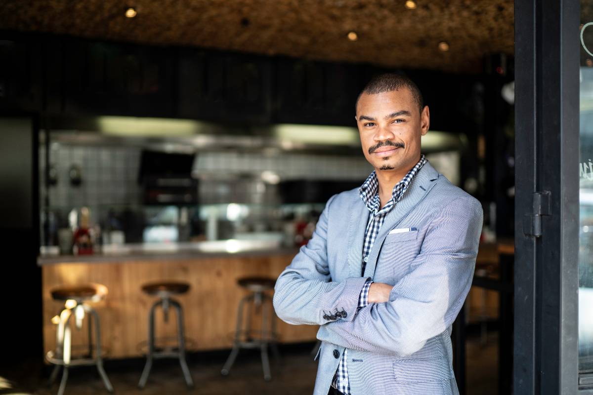 Proprietário de restaurante, de braços cruzados e posicionado na entrada de seu estabelecimento, transmitindo confiança e profissionalismo.