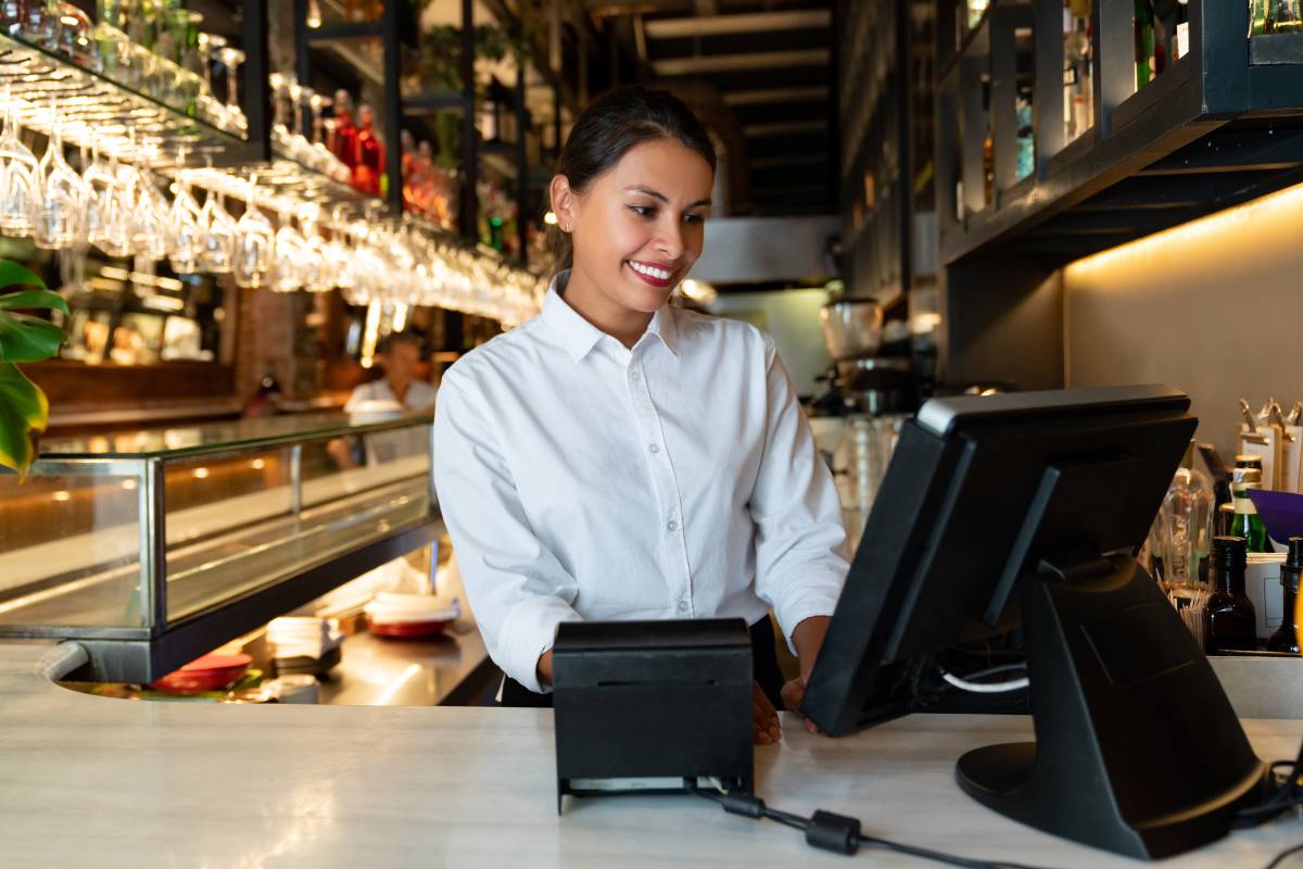 Funcionária sorridente usando o sistema de ponto de venda (PDV) no balcão de um restaurante, preparando-se para registrar um pedido.